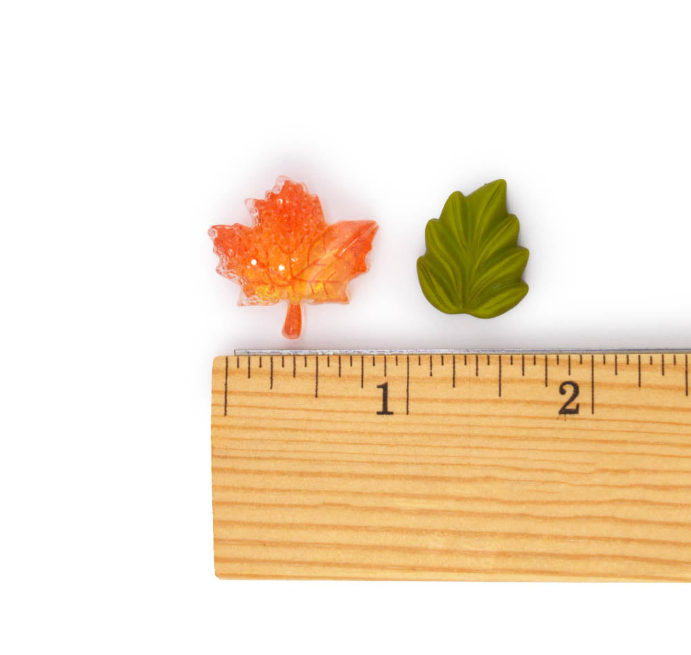 Two small leaf-shaped items, one orange maple leaf and one green oak leaf, shown above a wooden ruler on a white background to indicate size.