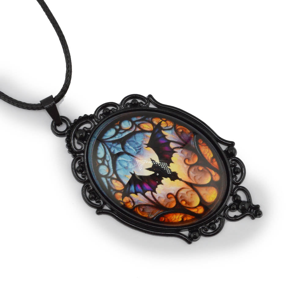 A gothic-style oval pendant necklace featuring a colorful stained-glass design of a bat and faces, set in an ornate black metal frame and suspended from a black cord, shown on a white background.