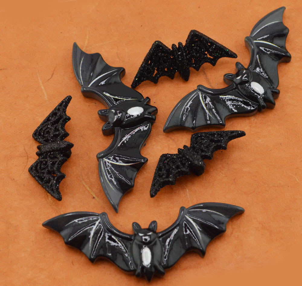Six black bat-shaped craft charms with glittery and glossy finishes, arranged on a textured orange background.