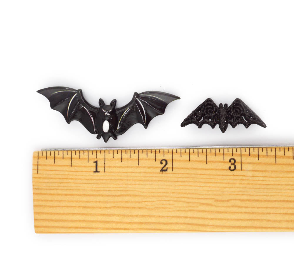 Two small black bat-shaped decorative pieces shown above a wooden ruler on a white background, with one larger bat and one smaller bat button