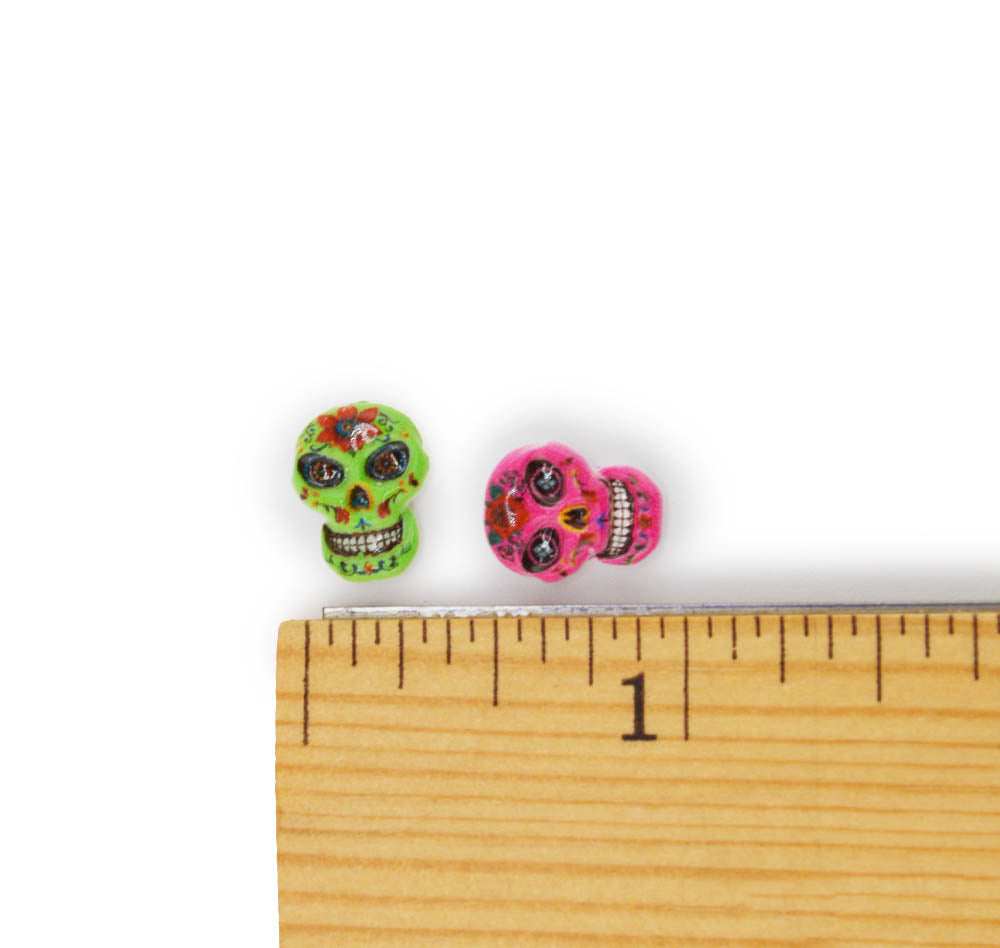 Two small sugar skull stud earrings shown next to a wooden ruler for scale on a white background. One earring is green with multicolor floral details, and the other is pink with black and white eye details.