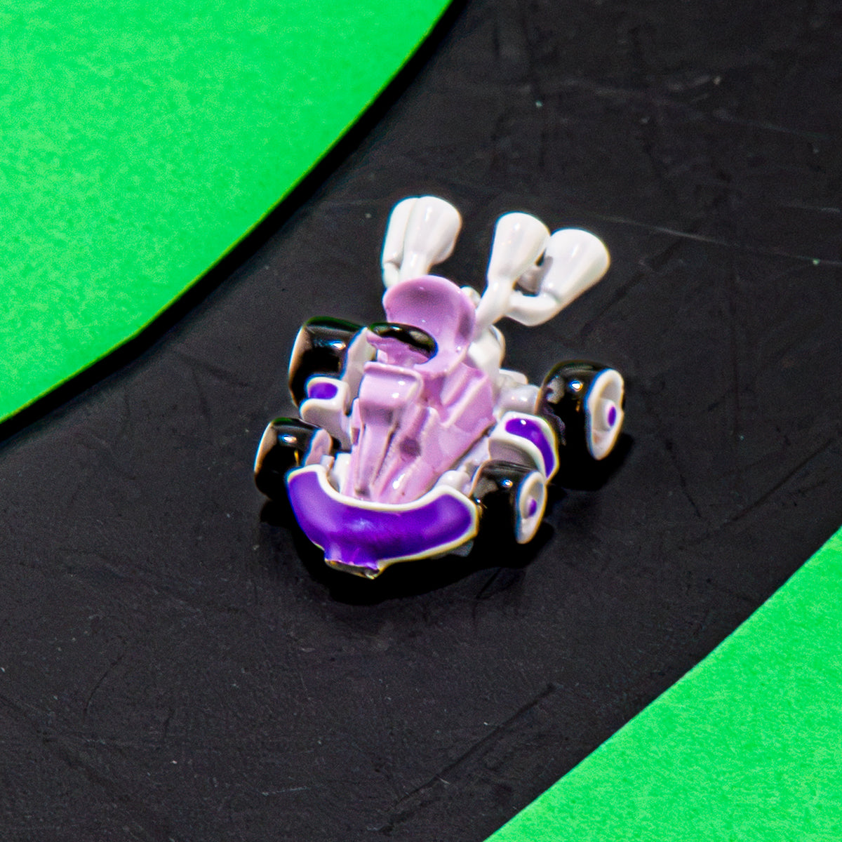 A small go-kart charm or pin featuring a purple and lavender body with white wheels and handlebars, set against a black textured background with bright green curved accents.