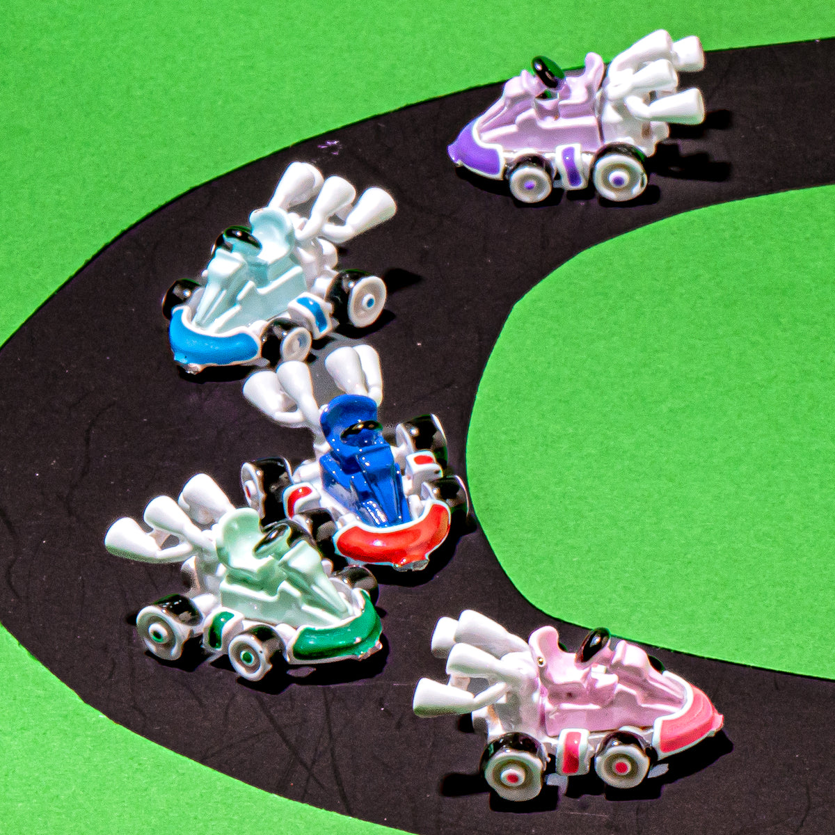 Five miniature toy race cars in assorted colors (white, blue, green, pink, purple) racing on a black curved track over a bright green background.