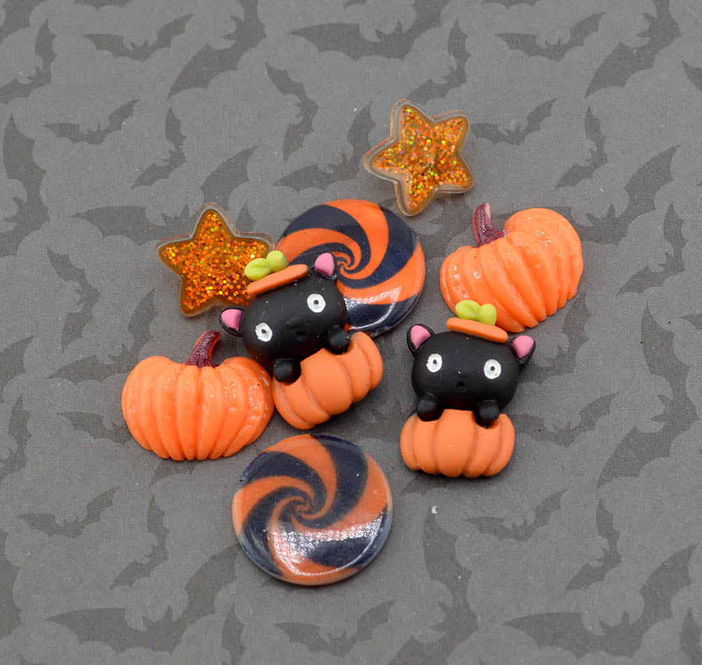 A set of Halloween-themed polymer clay charms and buttons featuring black cats in witch hats, orange pumpkins, a glittery orange star, and orange-and-black swirl designs, displayed on a gray background with a subtle bat pattern.
