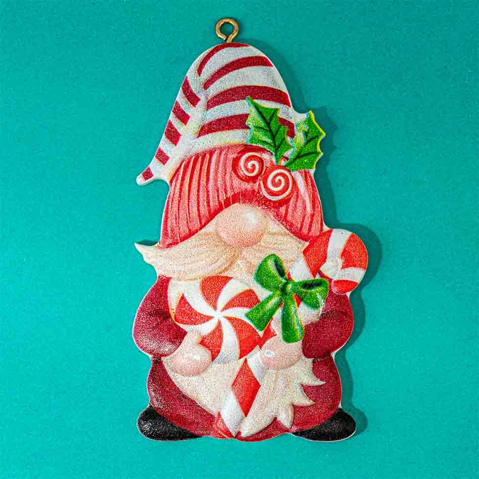 A festive Christmas gnome ornament featuring a red and white striped hat, red and white swirl patterns, green holly leaves, and candy cane motifs, shown against a solid teal background.