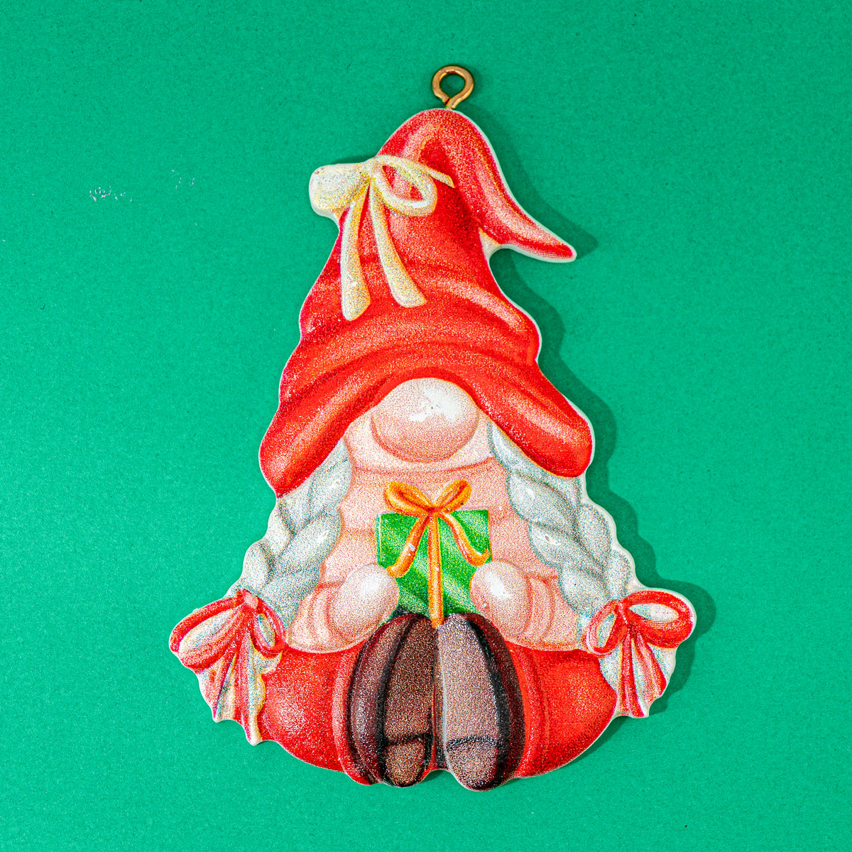 A Christmas gnome ornament featuring a red hat with a cream bow, white braided hair, a pink face, and red boots, holding a green gift box with an orange bow. The ornament is shown against a solid green background.