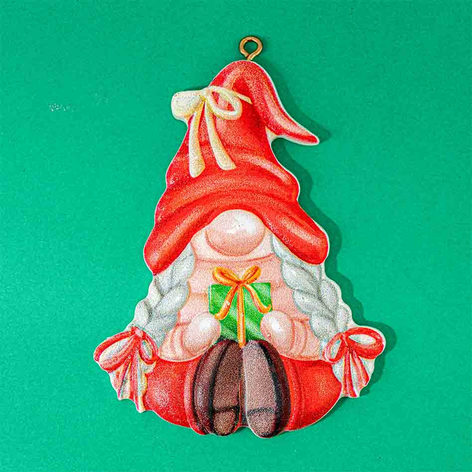 A Christmas gnome ornament featuring a red hat with a cream bow, white braided hair, a pink face, and red boots, holding a green gift box with an orange bow. The ornament is shown against a solid green background.