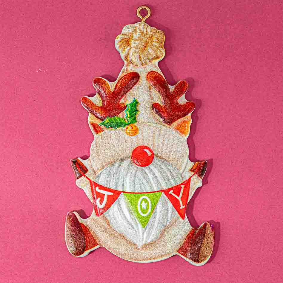 A vintage-style ceramic Christmas ornament shaped like a gnome with reindeer antlers, a red nose, and a 'JOY' banner, displayed on a solid pink background.