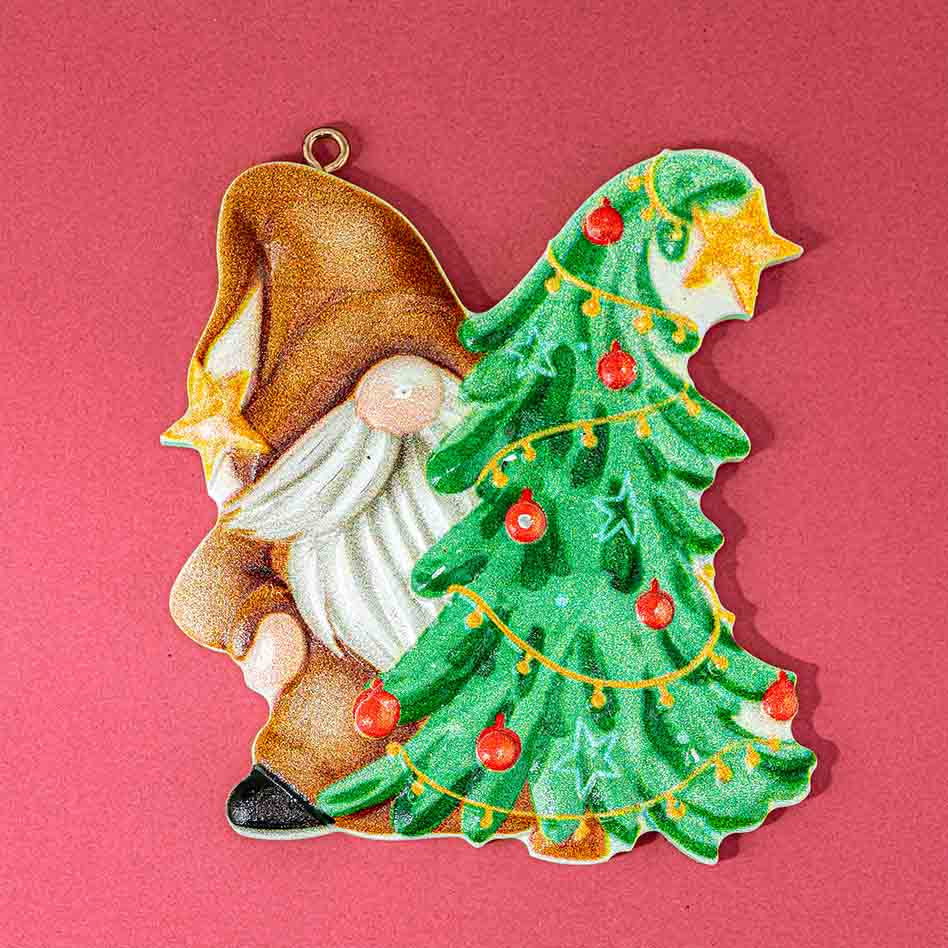 A Christmas ornament shaped like a gnome next to a decorated Christmas tree, shown on a solid pink background.
