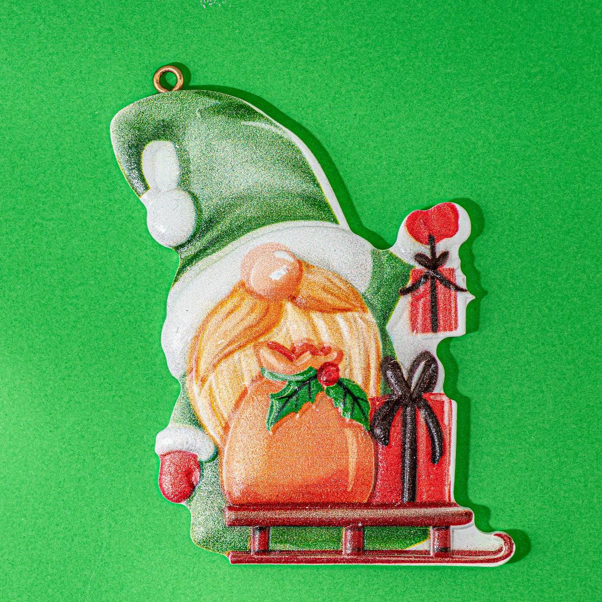 A Christmas gnome ornament featuring a green hat, white beard, orange beard, and a sleigh carrying red gifts, shown on a solid green background.