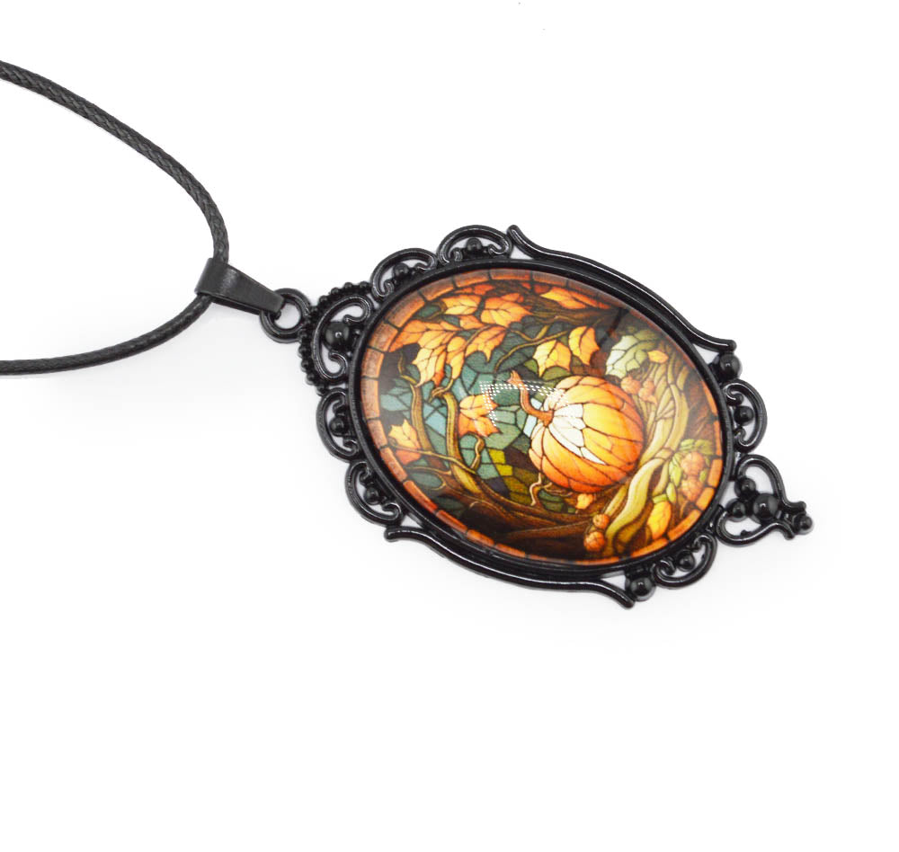 An oval pendant necklace featuring a colorful stained-glass style illustration of a pumpkin and autumn leaves, set in an ornate black metal frame and suspended from a black cord, shown on a plain white background.