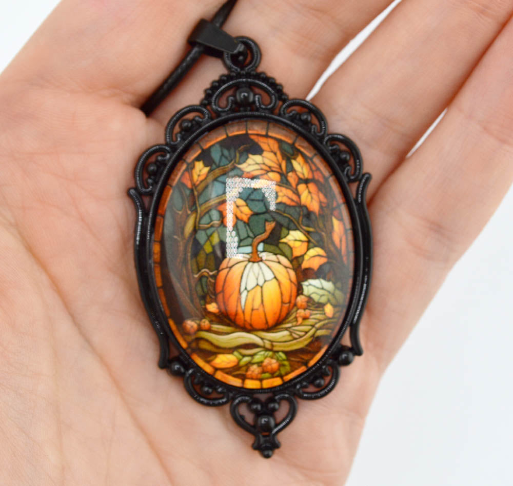 A hand holding an oval pendant necklace with a black ornate frame and a stained-glass-style autumn scene featuring a pumpkin, leaves, and a white letter 'O' in the center.