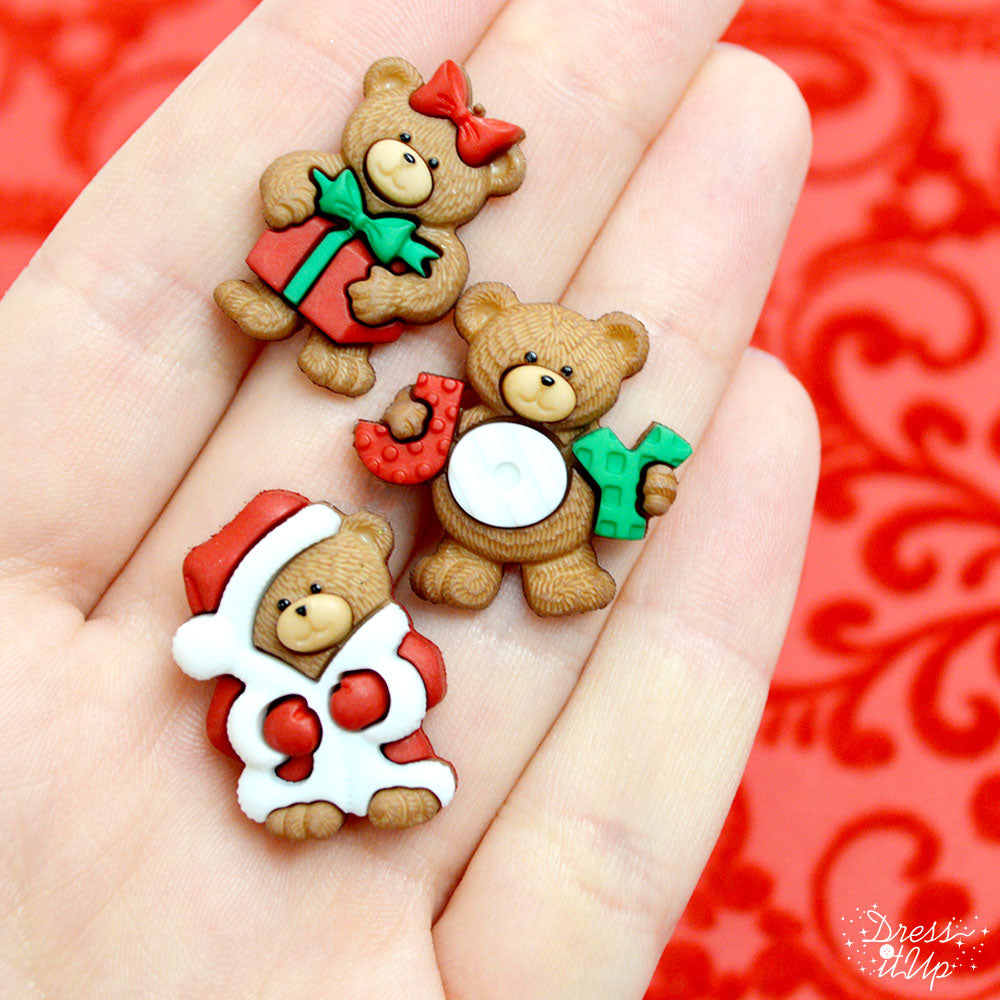 Three shank-back Christmas bear buttons under a Christmas tree with a Santa costume, a present, and a joy sign