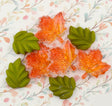 Six decorative craft embellishments featuring three translucent orange maple leaves with glitter and three solid green maple leaves, arranged on a floral-patterned background.