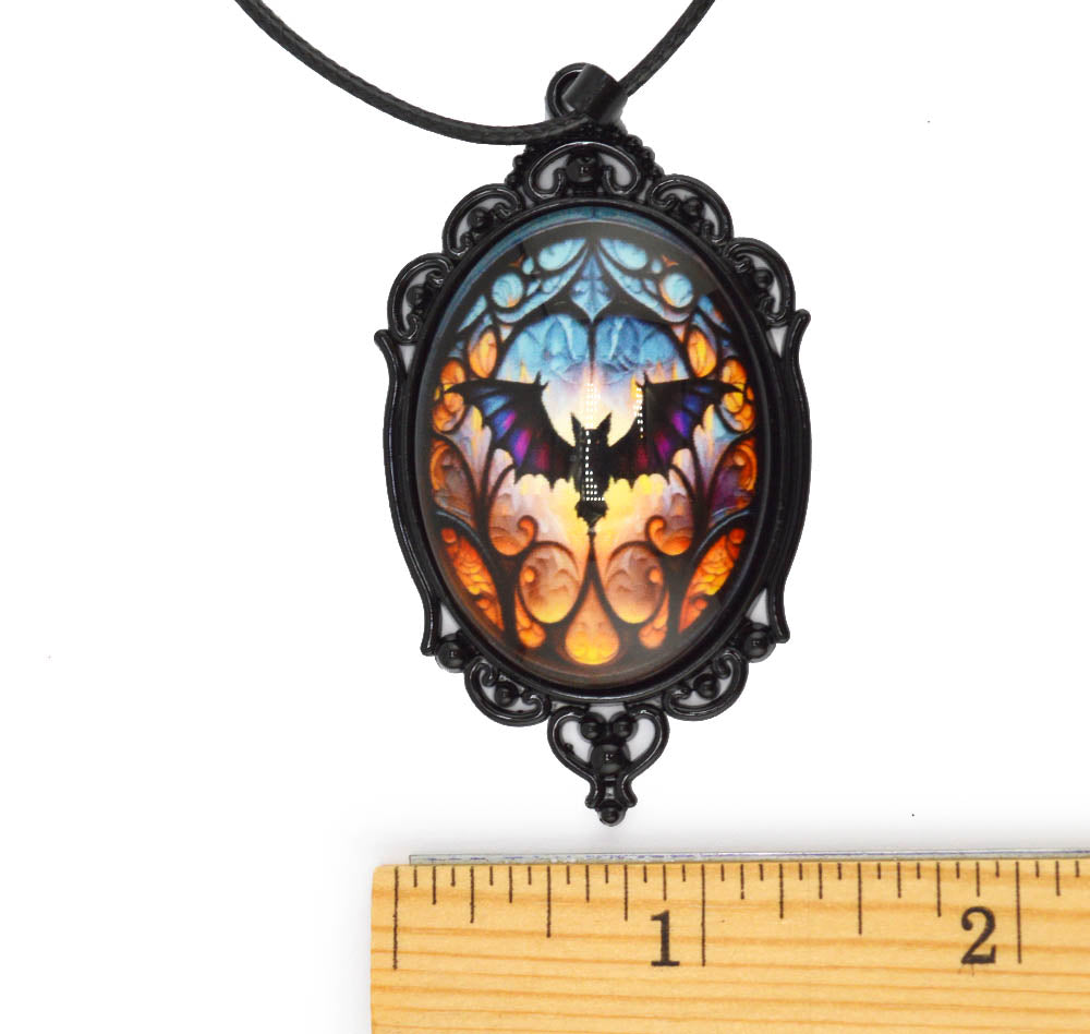 A gothic-style oval pendant necklace with a black filigree frame and a black cord, featuring a colorful stained-glass design with a bat motif. A wooden ruler is positioned below the pendant to show its scale against a white background.