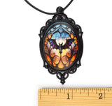 A gothic-style oval pendant necklace with a black filigree frame and a black cord, featuring a colorful stained-glass design with a bat motif. A wooden ruler is positioned below the pendant to show its scale against a white background.
