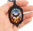 A hand holding an oval pendant necklace with a black ornate frame and a colorful stained-glass-style design featuring a bat silhouette in blue, purple, orange, and yellow.