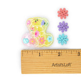 A small translucent teddy bear-shaped embellishment with pastel floral details is shown next to three flower-shaped pieces in coral, purple, and turquoise, with a wooden ruler labeled Artist'sLoft below for scale on a white background.