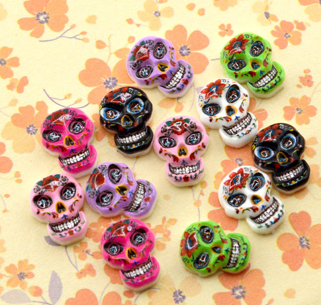 A group of colorful sugar skull cabochons in various designs and colors, including pink, purple, green, black, and white, arranged on a yellow floral background.