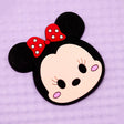 A Minnie Mouse Tsumtsum-style character plastic applique featuring a beige face, black ears and outline, and a red bow with white polka dots, shown on a light purple textured background.