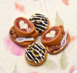 Three miniature ceramic coffee-themed charms on a floral paper background. Two are brown coffee cups with heart latte art on the inside, and one is a chocolate-draped pastry.