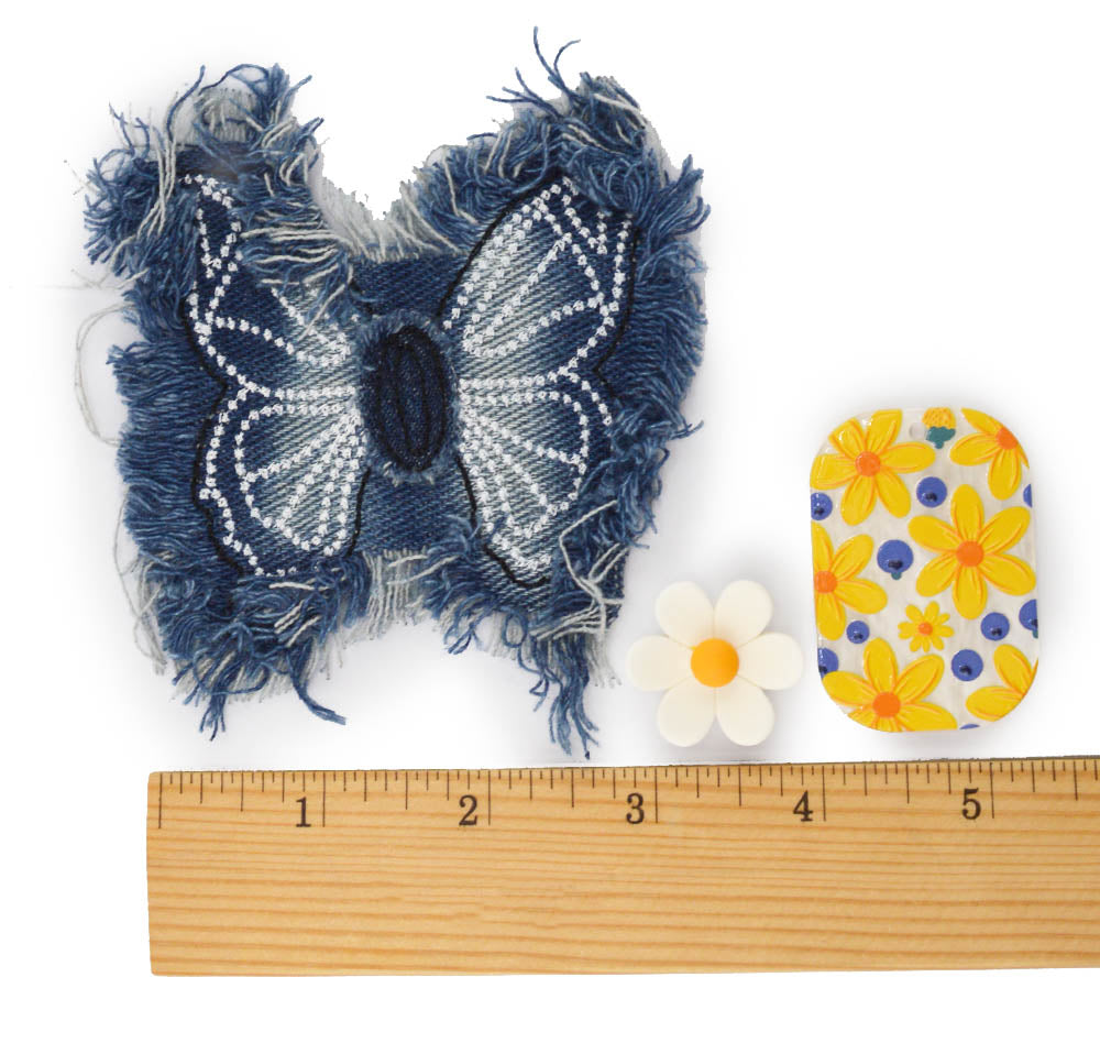 A handmade denim butterfly patch in blue and white, shown next to a wooden ruler, a small white daisy, and a yellow and blue floral patch on a white background.
