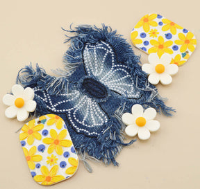 A decorative fabric patch featuring a blue denim-like background with a white beaded butterfly design and frayed edges, surrounded by four white daisy embellishments with yellow centers on a beige background.