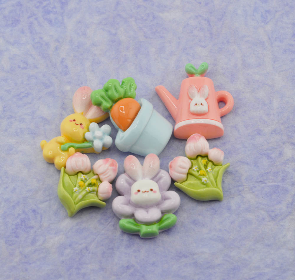 A set of cute pastel Easter-themed charms and embellishments, including a pink watering can with a bunny face, a yellow bunny holding a flower, a carrot in a pot, and pink tulips with a central bunny flower, arranged on a textured light purple background.
