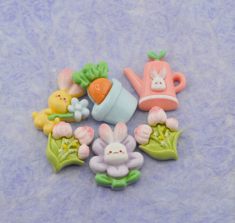 A set of cute pastel Easter-themed charms and embellishments, including a pink watering can with a bunny face, a yellow bunny holding a flower, a carrot in a pot, and pink tulips with a central bunny flower, arranged on a textured light purple background.