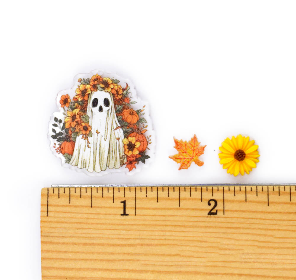 A Halloween-themed die-cut sticker featuring a white ghost surrounded by orange pumpkins, yellow flowers, and autumn leaves, displayed next to a wooden ruler and two small decorative pieces on a white background.