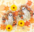 cute white ghost with a black face surrounded by orange autumn flowers, pumpkins, and leaves, displayed on a floral-patterned cream background with real yellow flowers and fall leaves.
