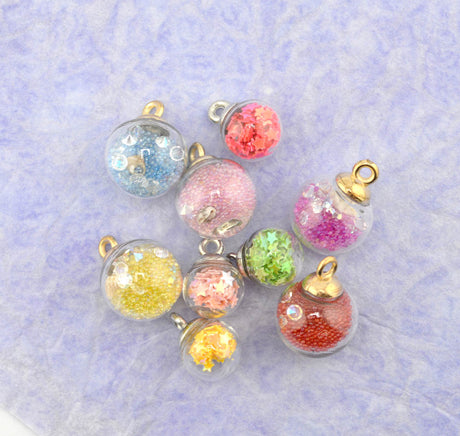 Seven small clear glass orb charms filled with colorful glitter in pink, blue, yellow, green, red, and orange, each with a gold-tone loop at the top, arranged on a light lavender textured background.