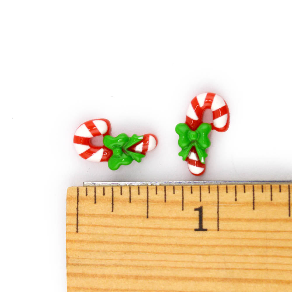 Two small candy cane resin Christmas-themed charms shown next to a wooden ruler for scale on a white background.