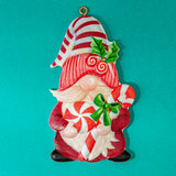 A festive Christmas gnome ornament featuring a red and white striped hat, red and white swirl patterns, green holly leaves, and candy cane motifs, shown against a solid teal background.