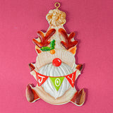 A vintage-style ceramic Christmas ornament shaped like a gnome with reindeer antlers, a red nose, and a 'JOY' banner, displayed on a solid pink background.