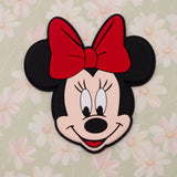 A Minnie Mouse head-shaped product, likely a patch, sticker, or small plaque, featuring her iconic red bow, black ears, and pink face against a light green floral background.