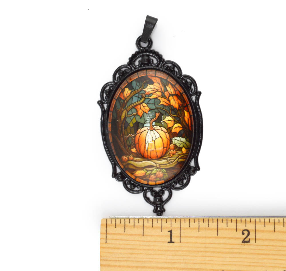 An oval pendant with an ornate black filigree frame and a stained-glass-style autumn scene featuring a pumpkin, leaves, and birds, shown above a wooden ruler for scale on a white background.