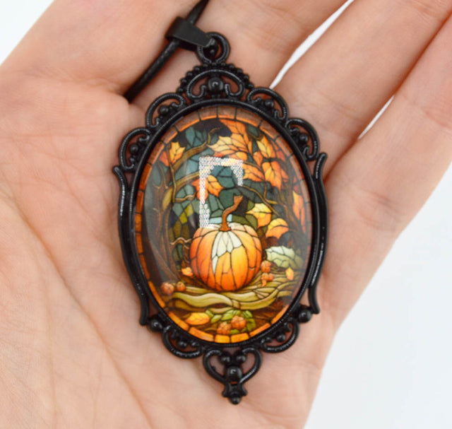 A hand holding an oval pendant necklace with a black ornate frame and a stained-glass-style autumn scene featuring a pumpkin, leaves, and a white letter 'O' in the center.