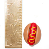 A small hot dog-shaped item with a tan bun, red filling, and yellow mustard graphic, shown next to a metal ruler on a white background.