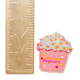 A small pink popcorn bucket charm or brooch with white popcorn and orange kernels, shown next to a gold-colored ruler for scale on a white background.