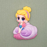 A glossy cartoon-style craft embellishments of a girl with yellow hair in a bun, wearing a pink and purple dress, seated on a purple cloud-like base against a plain green background.