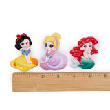 Three colorful fairy tale princess figurines (Snow White, Cinderella, and Ariel) displayed side-by-side above a wooden ruler on a white background.
