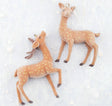 Two flocked deer charms in a warm tan color with small cream spots, shown on a light textured background. One deer has antlers and the other does not, both have small hanging loops at the top of their heads.