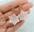 A pair of handmade snowflake dangle earrings held in a hand. The earrings feature gold-plated snowflake studs with tiny clear crystals and large, intricate snowflake drops made of white seed beads with clear rhinestones and a central pearl.