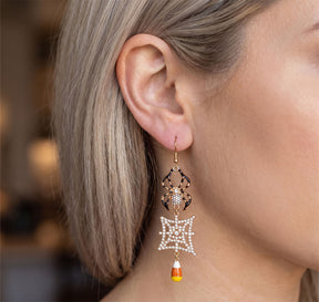 A close-up of a woman's ear wearing a dangling Halloween earring with a gold spider, a white pearl web, and a small candy corn charm.