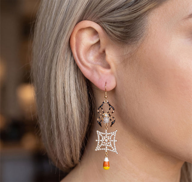 A close-up of a woman's ear wearing a dangling Halloween earring with a gold spider, a white pearl web, and a small candy corn charm.