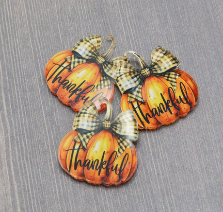 Three decorative autumn pumpkins plastic charms arranged on a gray wood-textured background. Each pumpkin is orange with the word "Thankful" written in black script and topped with a beige and black plaid bow.