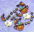 Halloween-themed decorative charms and letter beads arranged on a crinkled light blue background. The set includes two orange jack-o'-lantern teacups with colorful ghost figures wearing witch hats, two white smiling ghosts, two purple high-heeled witch shoes, and round black beads spelling TRICK OR TREAT with gold letters.