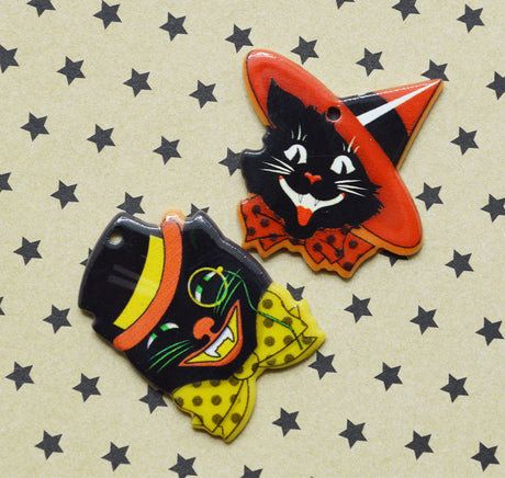 Two Halloween-themed decorative charms shaped like a black cat in a witch hat and a black cat wearing a bow tie, displayed on a beige background with a black star pattern.