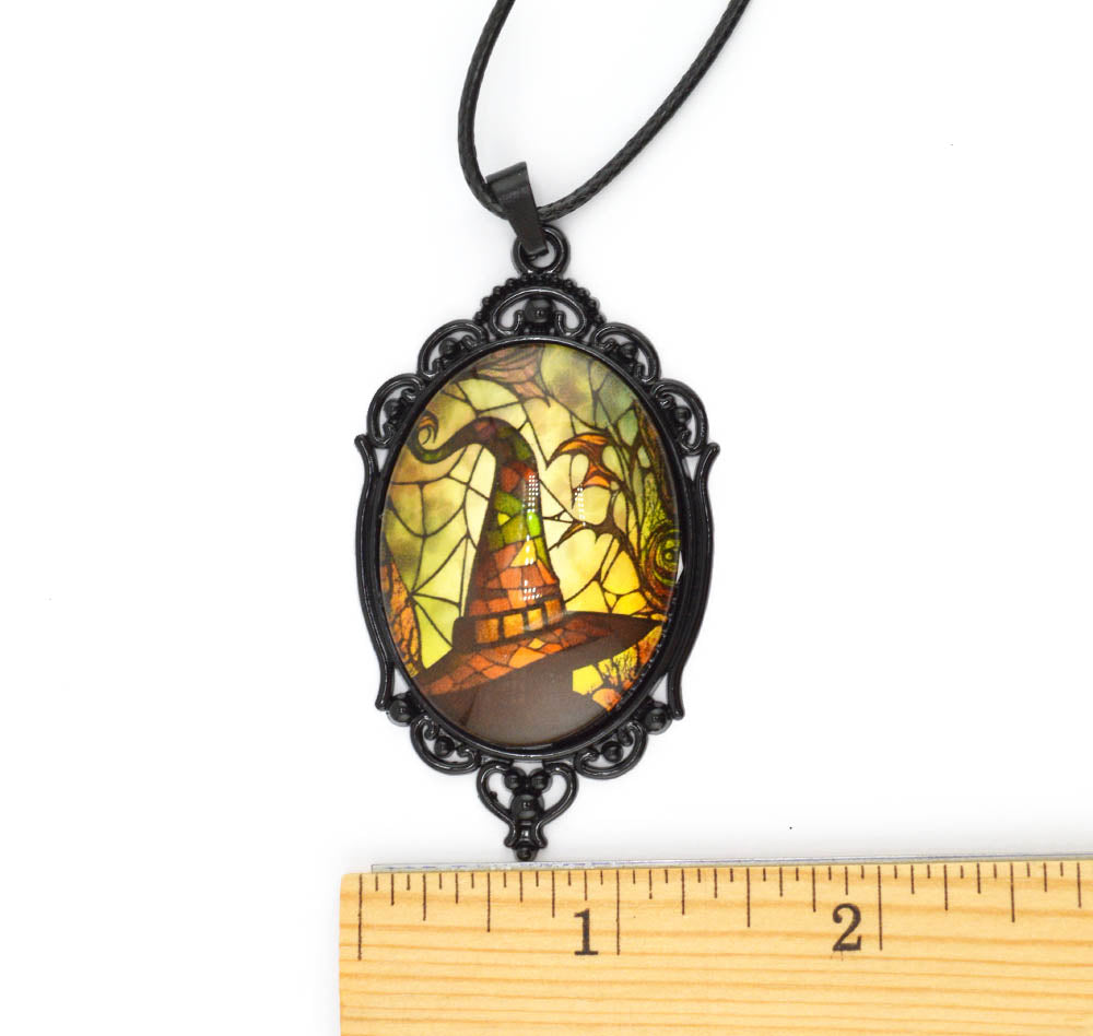 A gothic-style oval pendant necklace with a black ornate frame and a stained-glass window design featuring a witch's hat, spiderwebs, and a bat. A wooden ruler is placed below the pendant to show its scale against a white background.